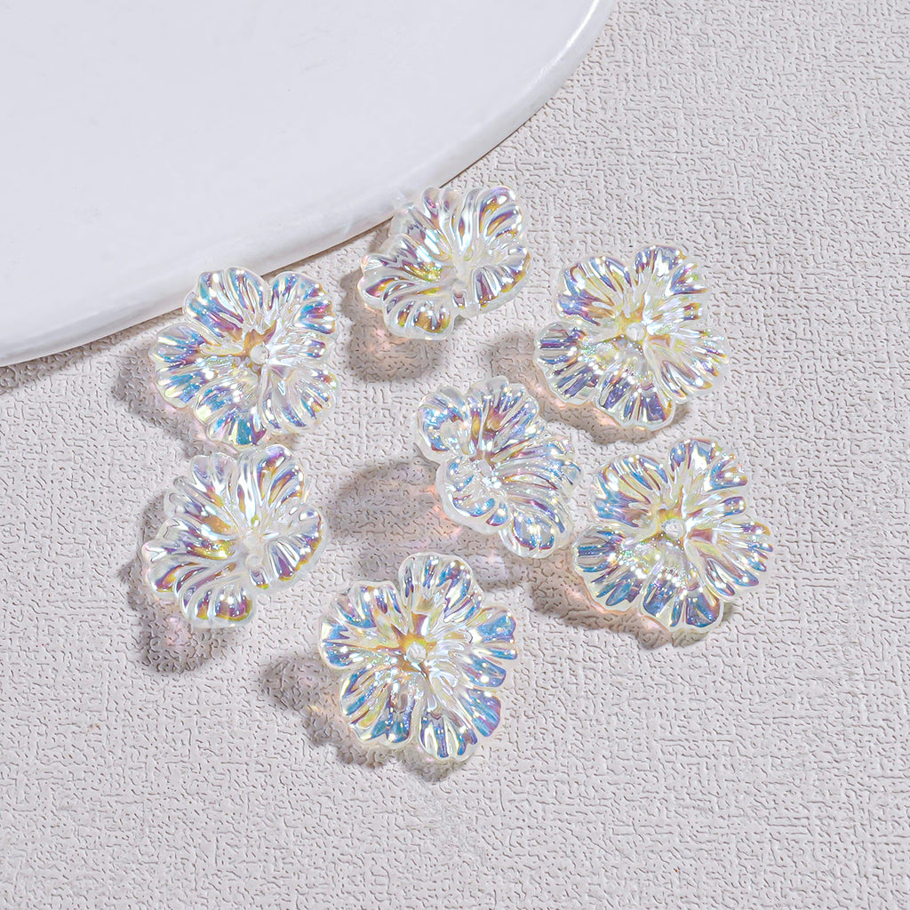 Eight iridescent clear resin flower beads arranged on a textured gray surface by a white plate edge