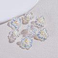 Eight iridescent clear resin flower beads arranged on a textured gray surface by a white plate edge