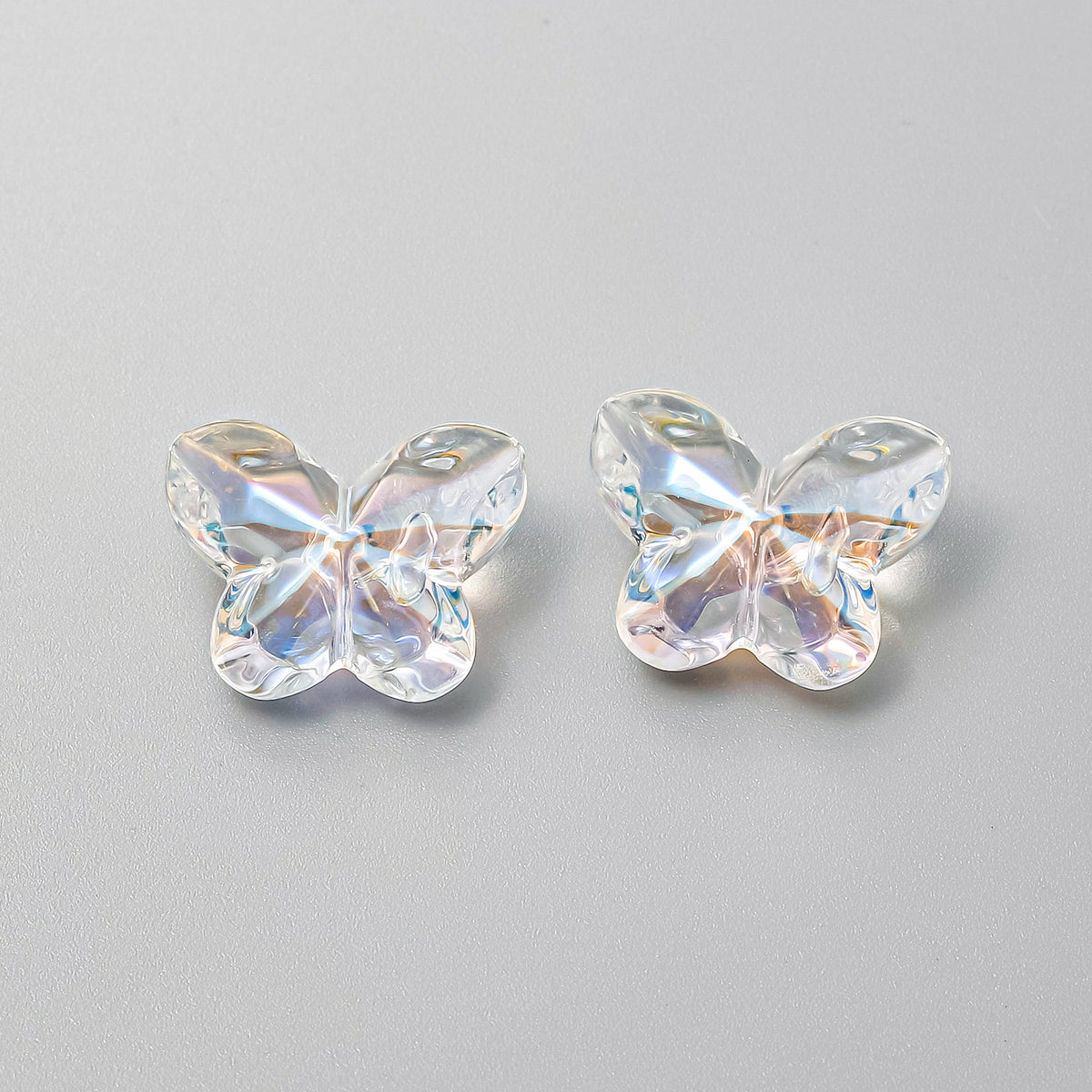 Two clear faceted butterfly-shaped beads with iridescent finish, side by side on a neutral gray background