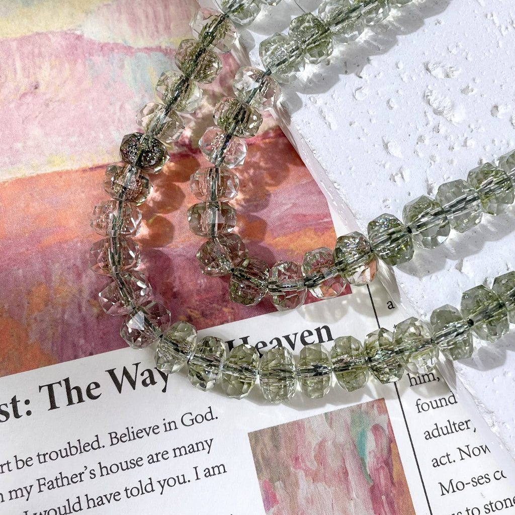 Strands of faceted clear beads with a light olive-green tint on a white textured block and colorful printed page