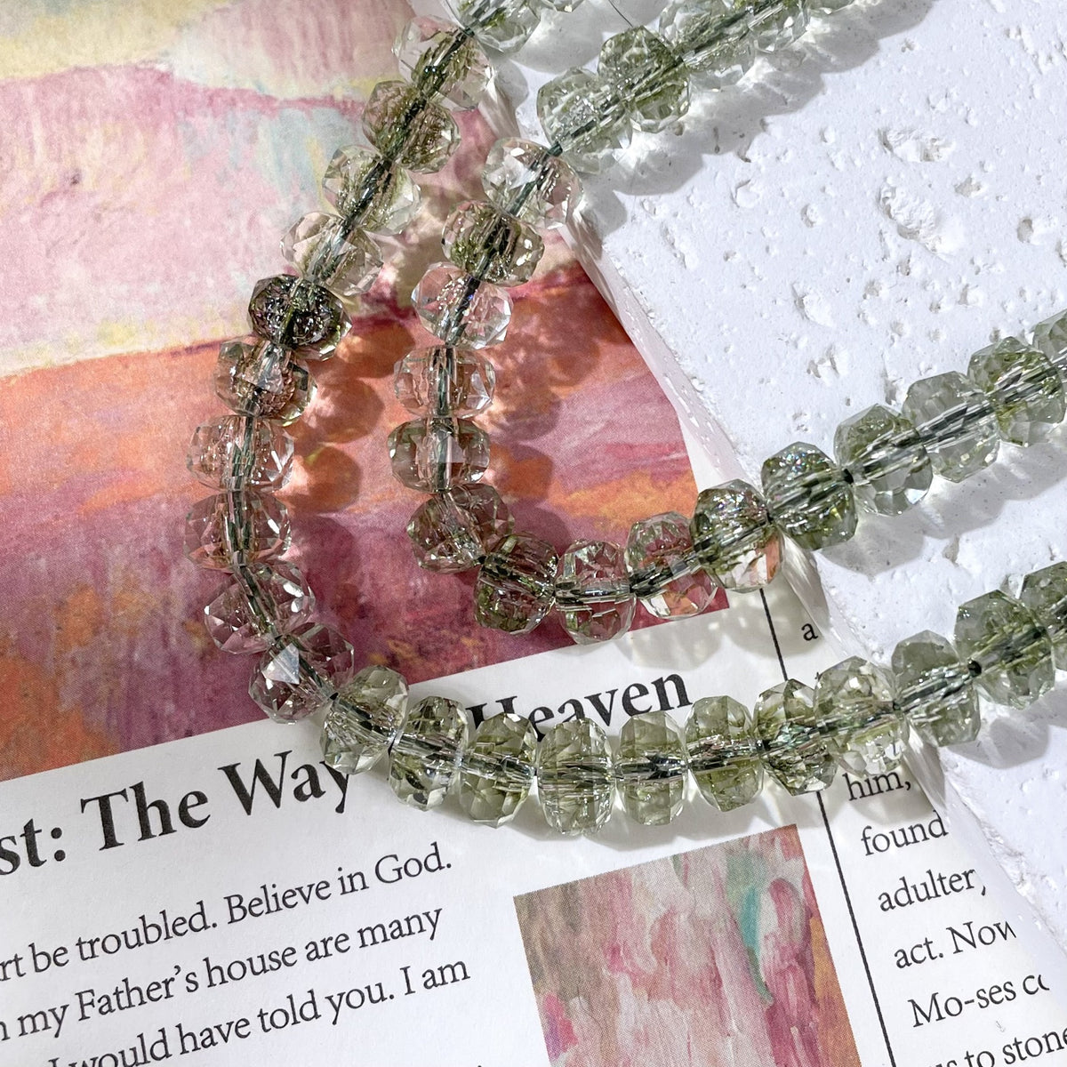 Strands of faceted clear beads with a light olive-green tint on a white textured block and colorful printed page