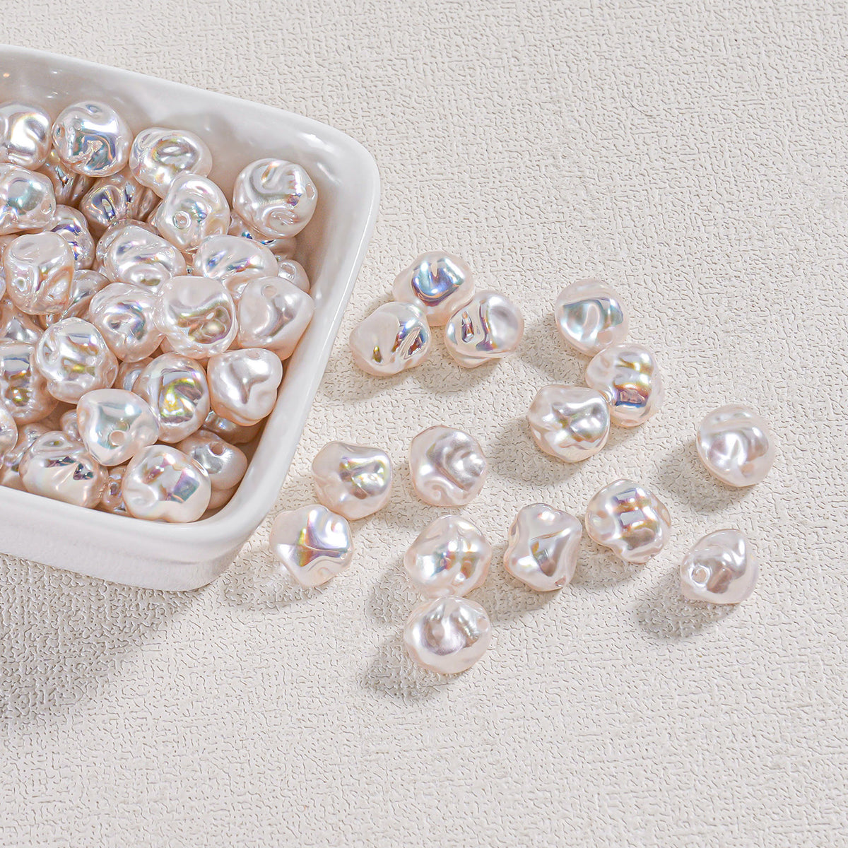 Iridescent baroque faux pearl beads in a white ceramic bowl with several scattered on a textured surface