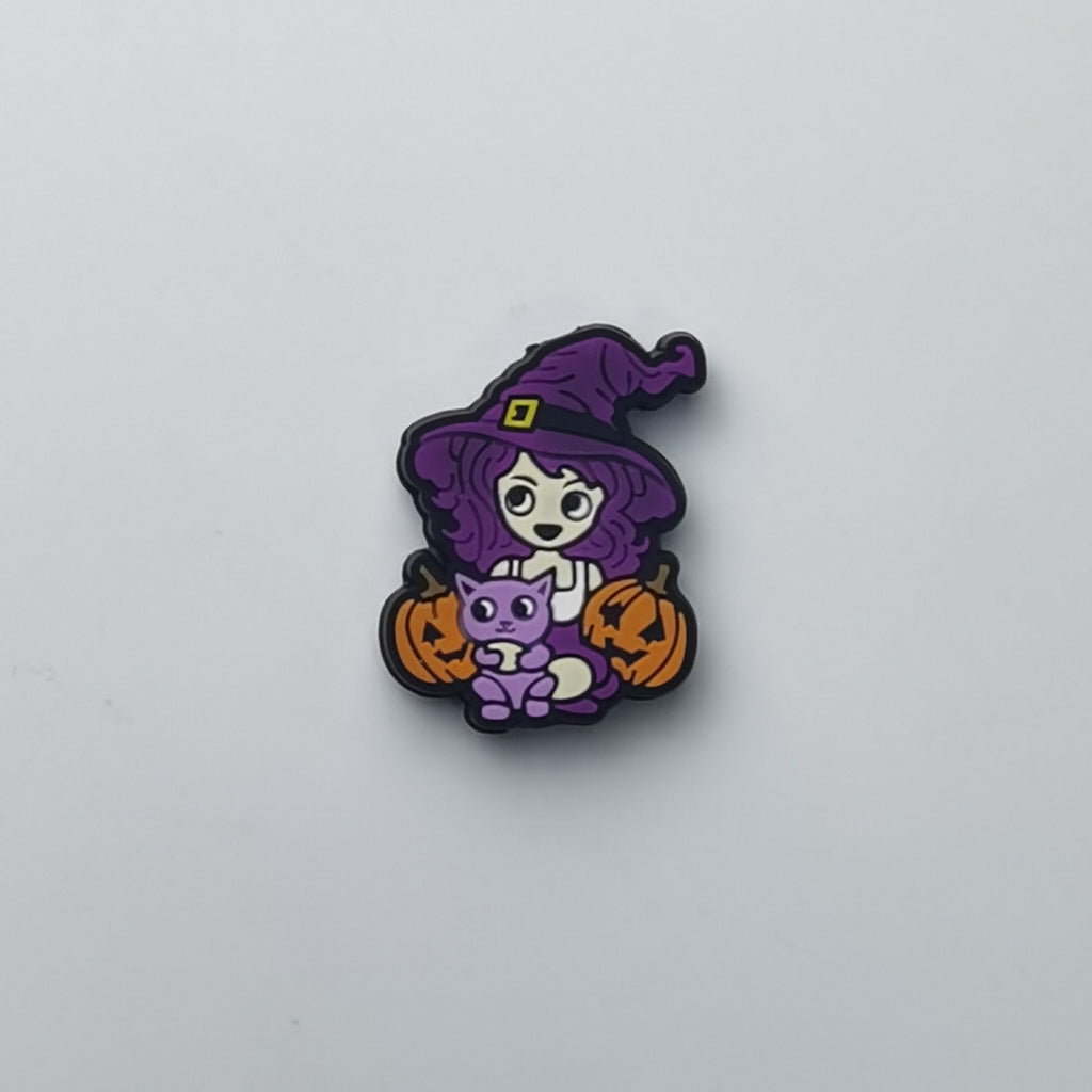 A small Halloween-themed charm or pin featuring a cartoon witch with a purple hat and hair, holding a purple cat and orange pumpkins, shown on a plain light background.