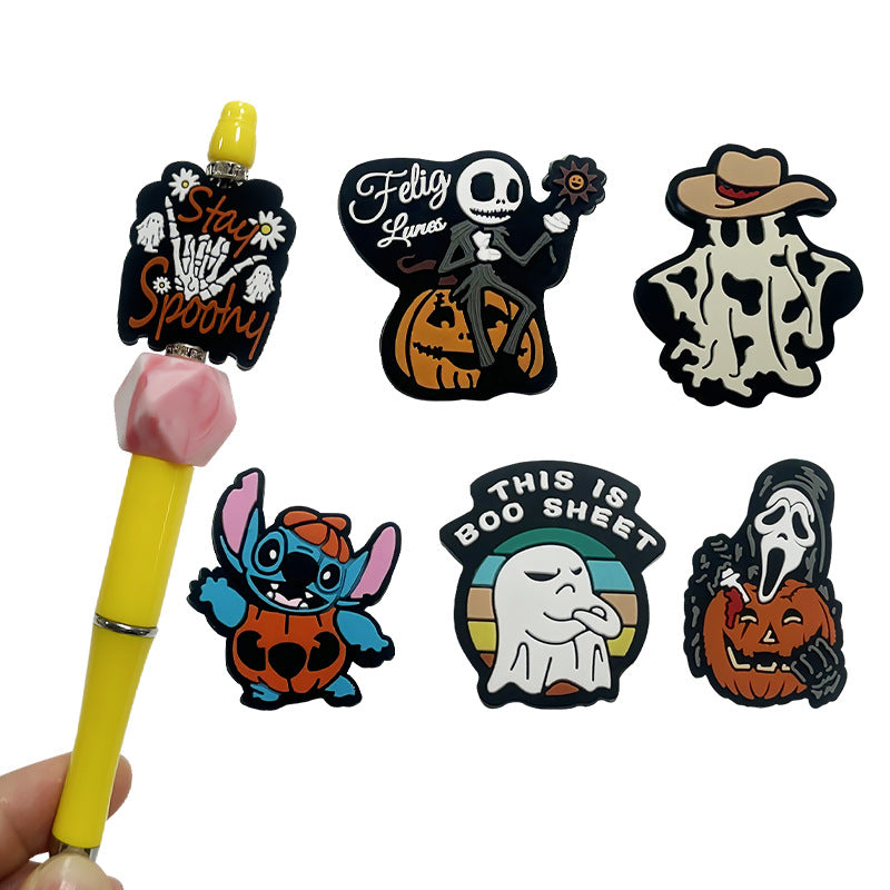 Six colorful Halloween silicone beads with skeleton, ghost, and pumpkin designs beside yellow pen topped with pink bead.