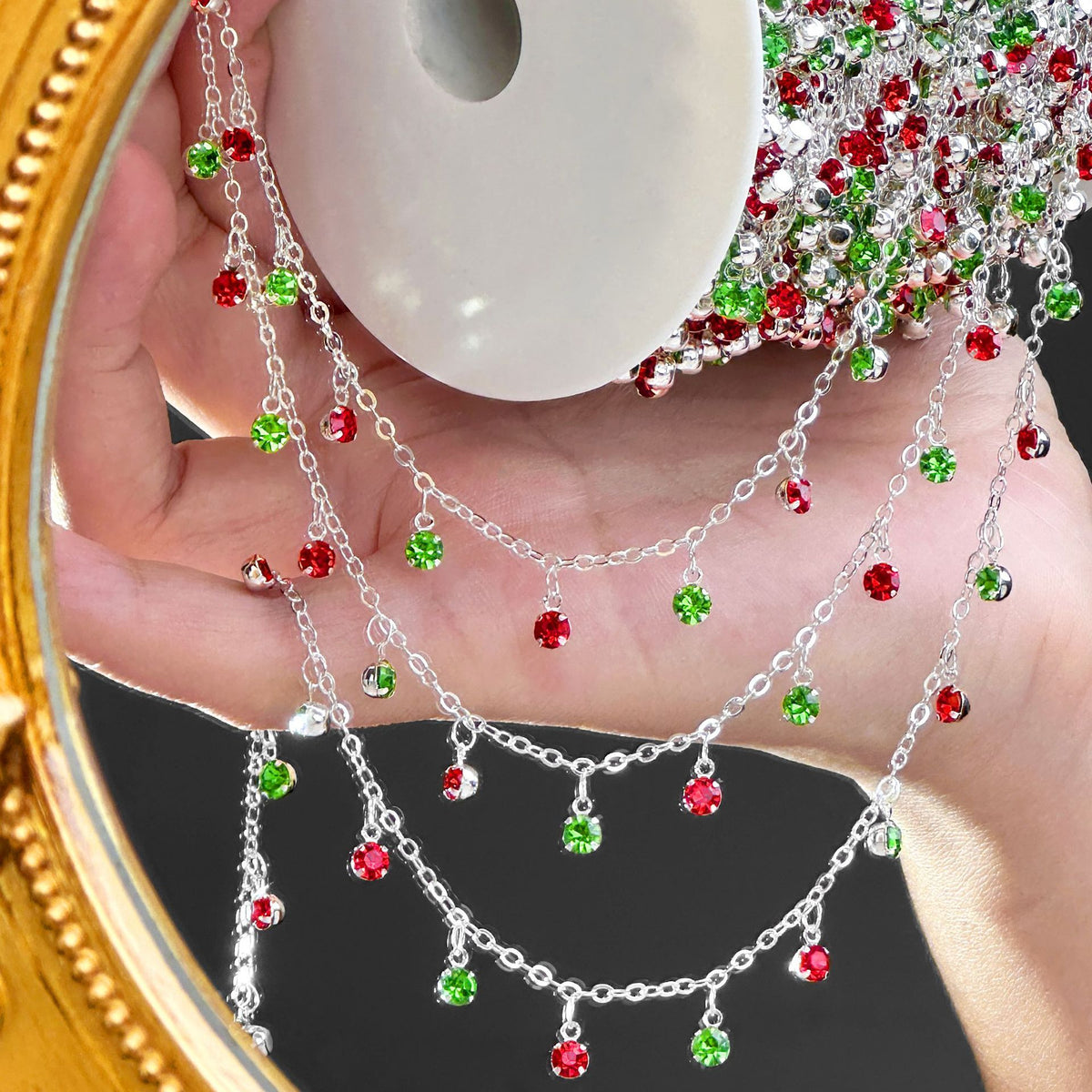 A hand holds a silver holiday garland with dangling chains and red and green crystal ornaments, with a gold mirror frame partially visible.