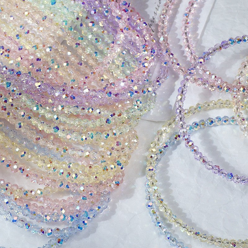 A close-up of multiple strands of iridescent beaded chains or cords arranged on a white textured surface. The beads reflect pastel rainbow colors including pink, purple, blue, yellow, and green.