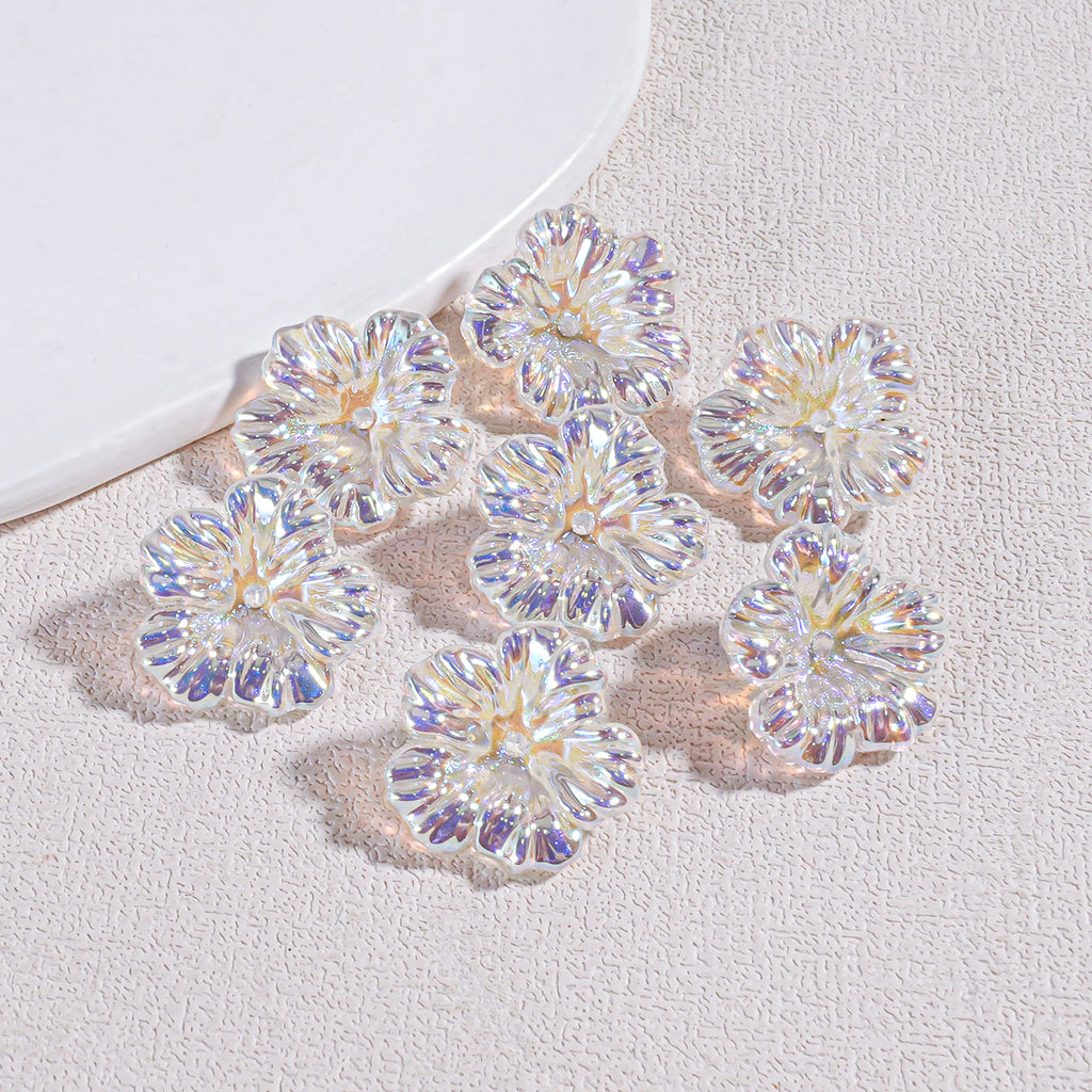 Nine iridescent holographic resin flower beads on a textured beige surface by a white curved prop