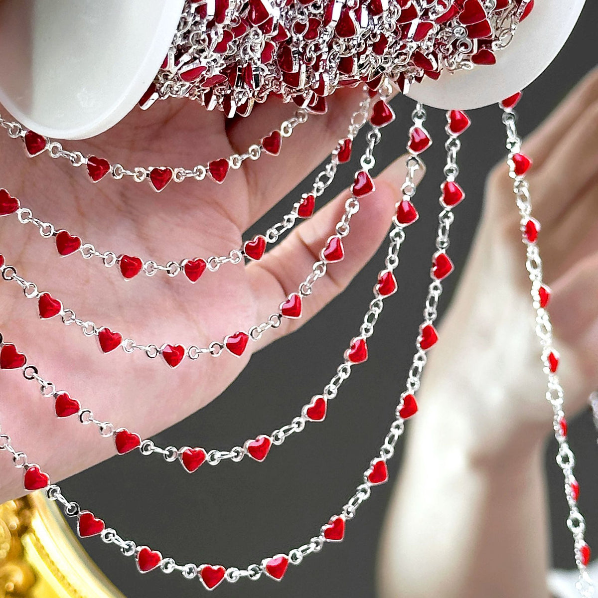 Hand holding silver-tone chain strands with red enamel heart beads, spool visible at top
