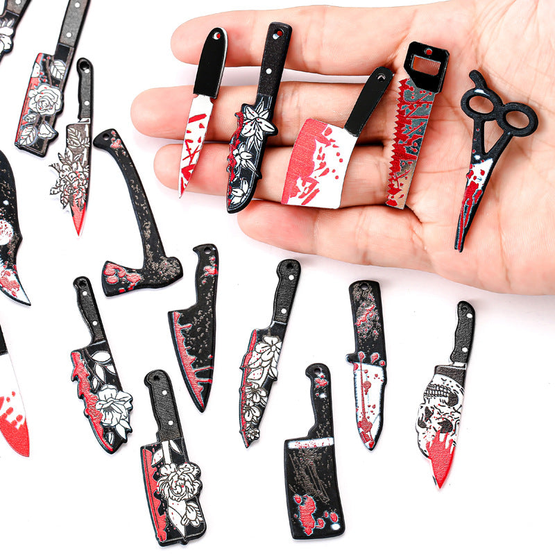 A hand holds several miniature toy kitchen knives and scissors with black handles, white floral patterns, and red blood splatter details, with more of the same scattered on a white background.