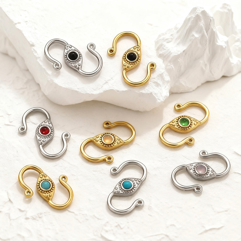 Eight stainless steel S-hook clasps, silver and gold finishes with colorful oval center stones, on a white surface