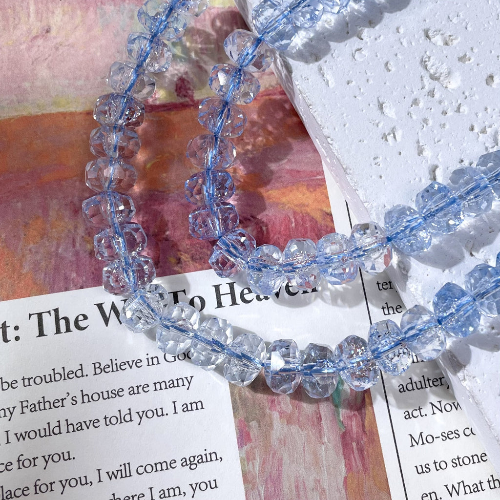 Light blue faceted crystal beads strung on a cord on a printed book page and a textured white stone slab