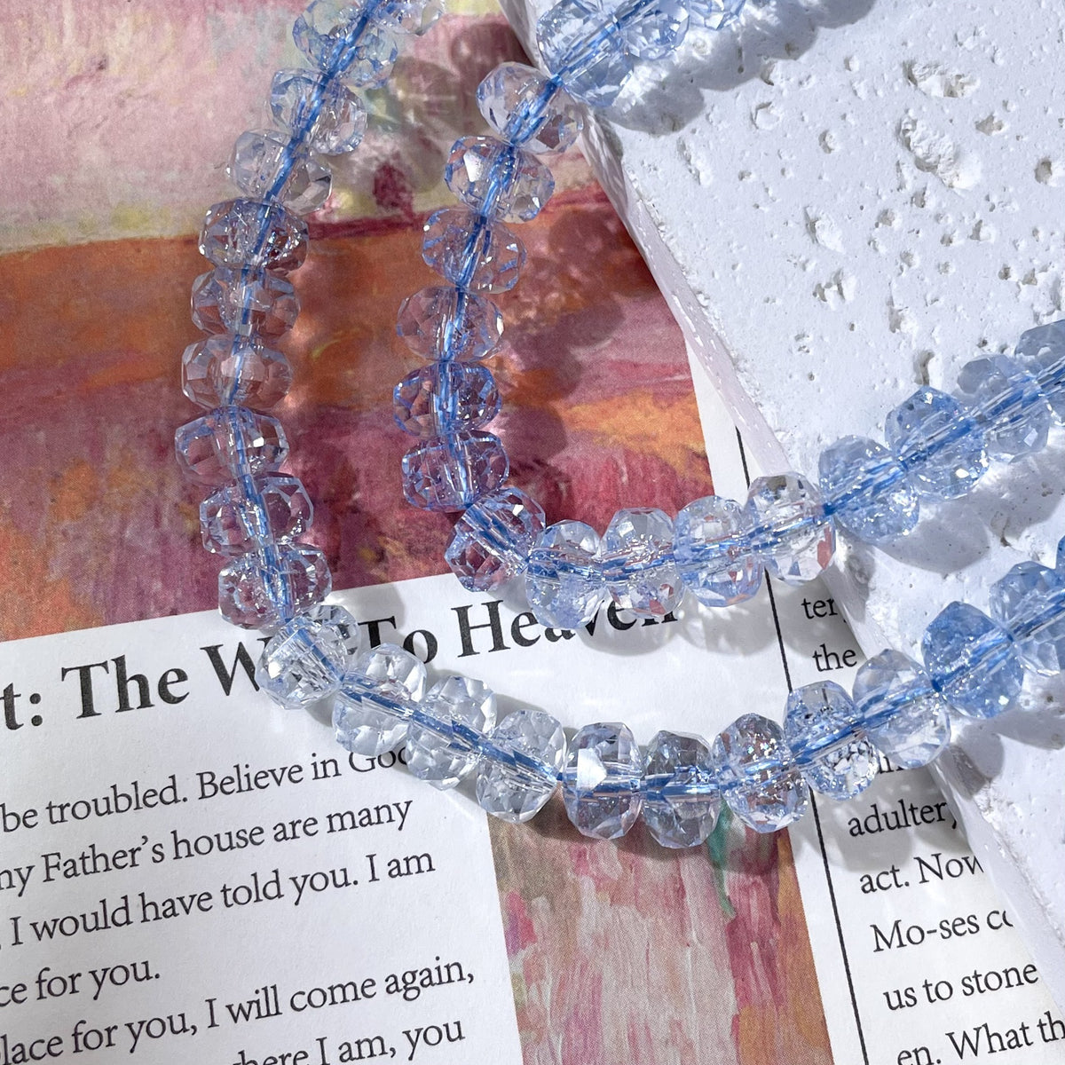 Light blue faceted crystal beads strung on a cord on a printed book page and a textured white stone slab