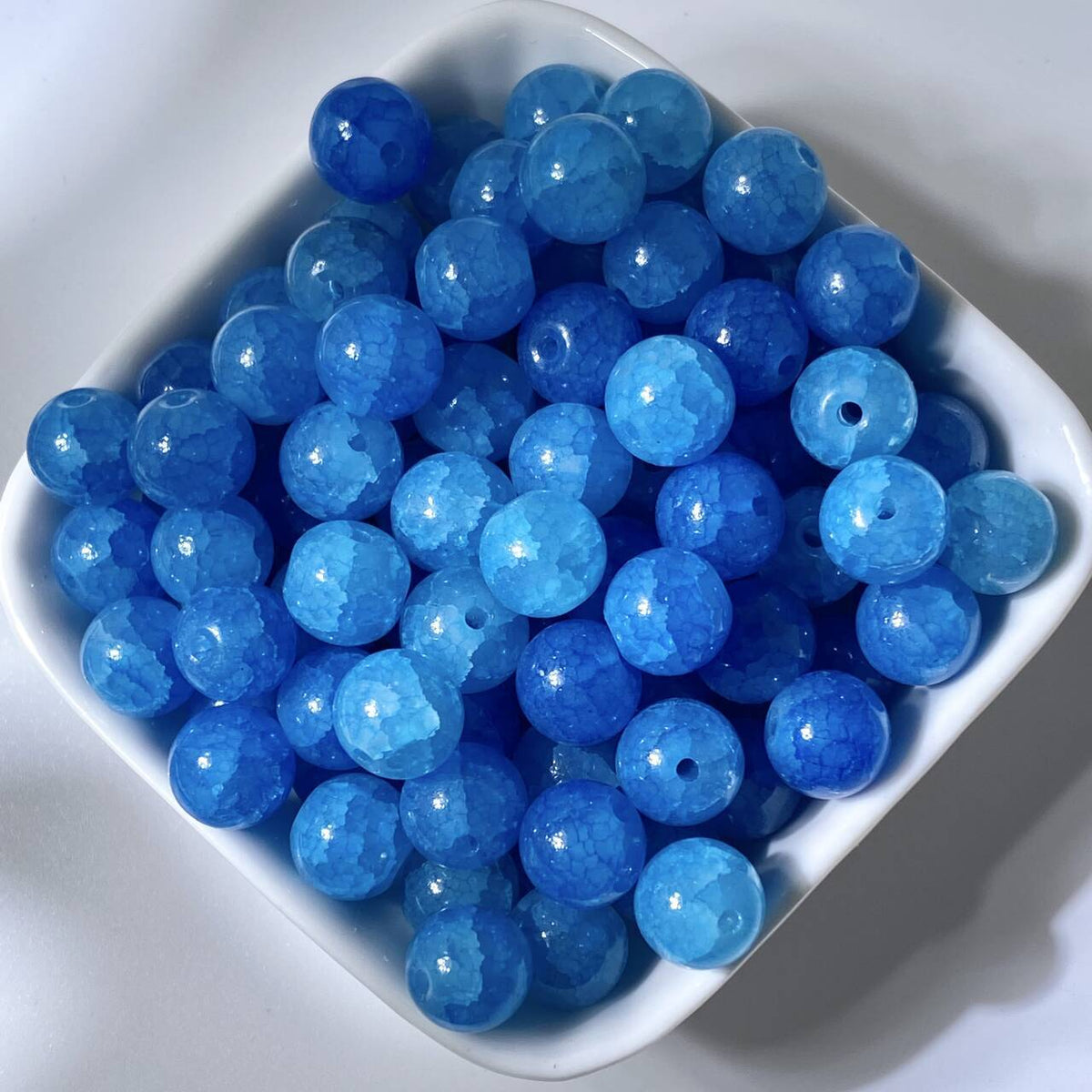 Blue 10mm water-dyed jade-style crackle glass beads with holes in a white square dish
