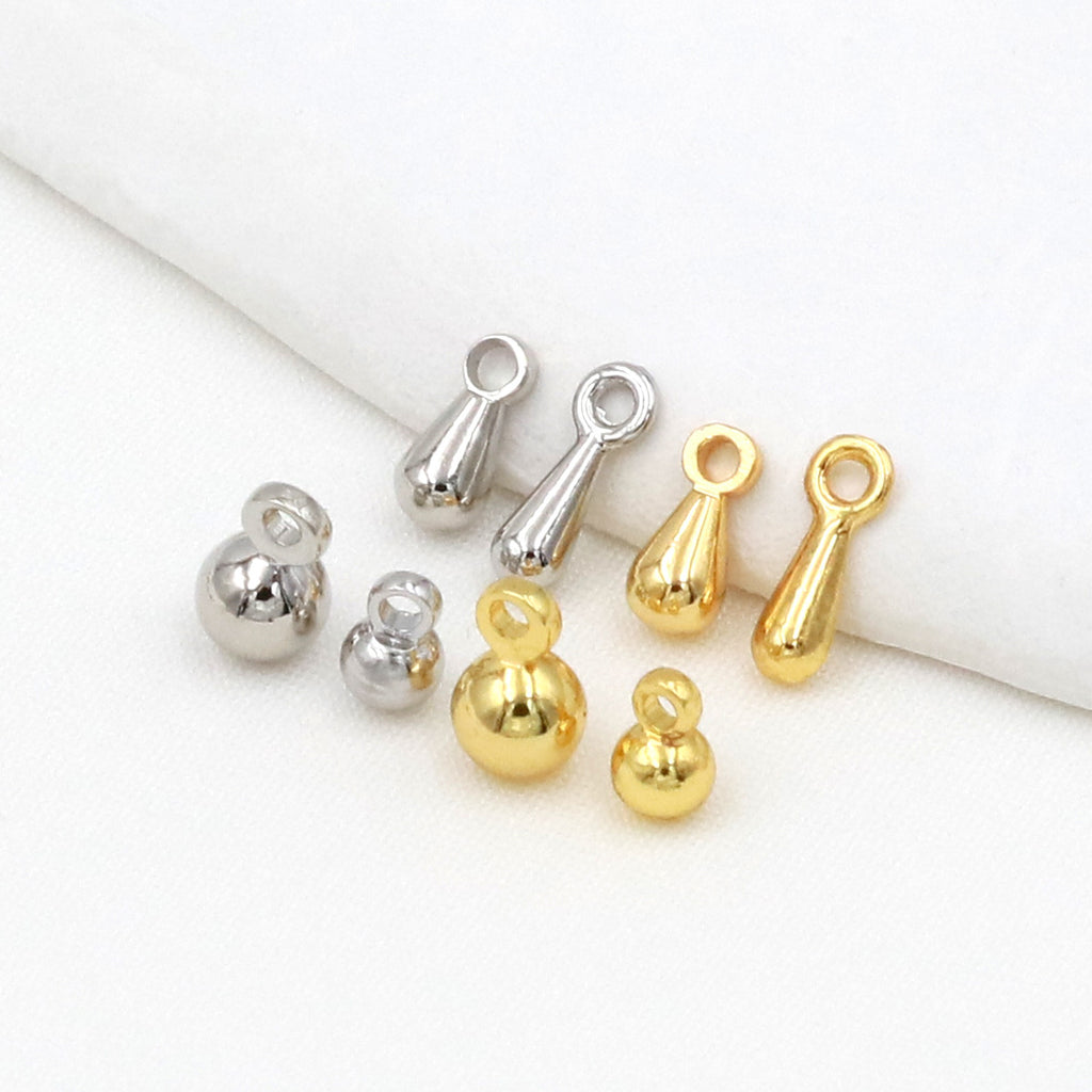 Six gold and silver ball, teardrop, and elongated drop charms arranged on a white fabric surface