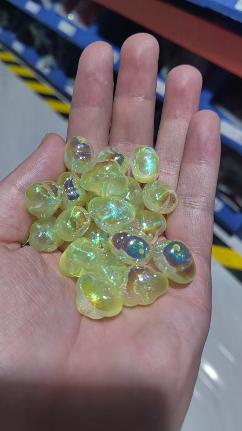 A hand holding small translucent iridescent yellow-green cloud-shaped resin beads with rainbow reflections