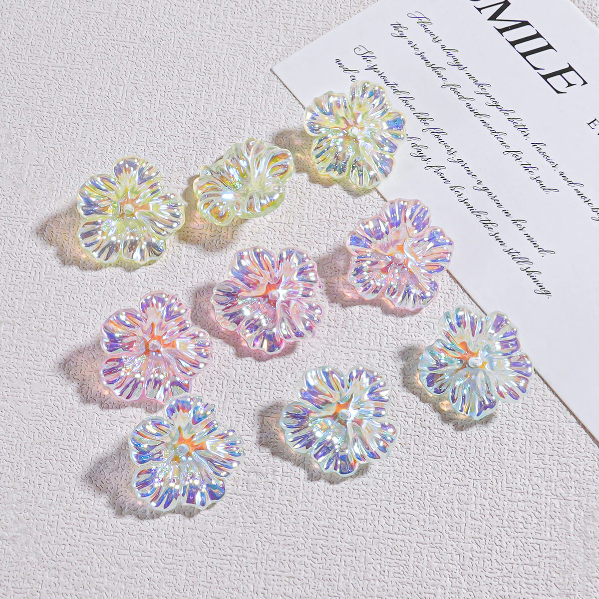 Nine iridescent holographic resin flower beads arranged on a textured white surface by a card