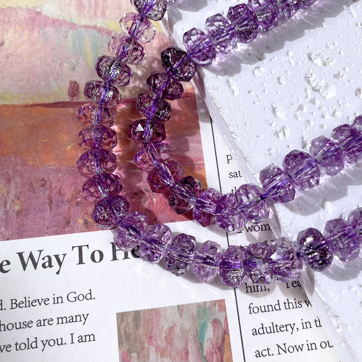Purple faceted glass beads with a glittery sparkle arranged in loops over a printed magazine page and textured white paper
