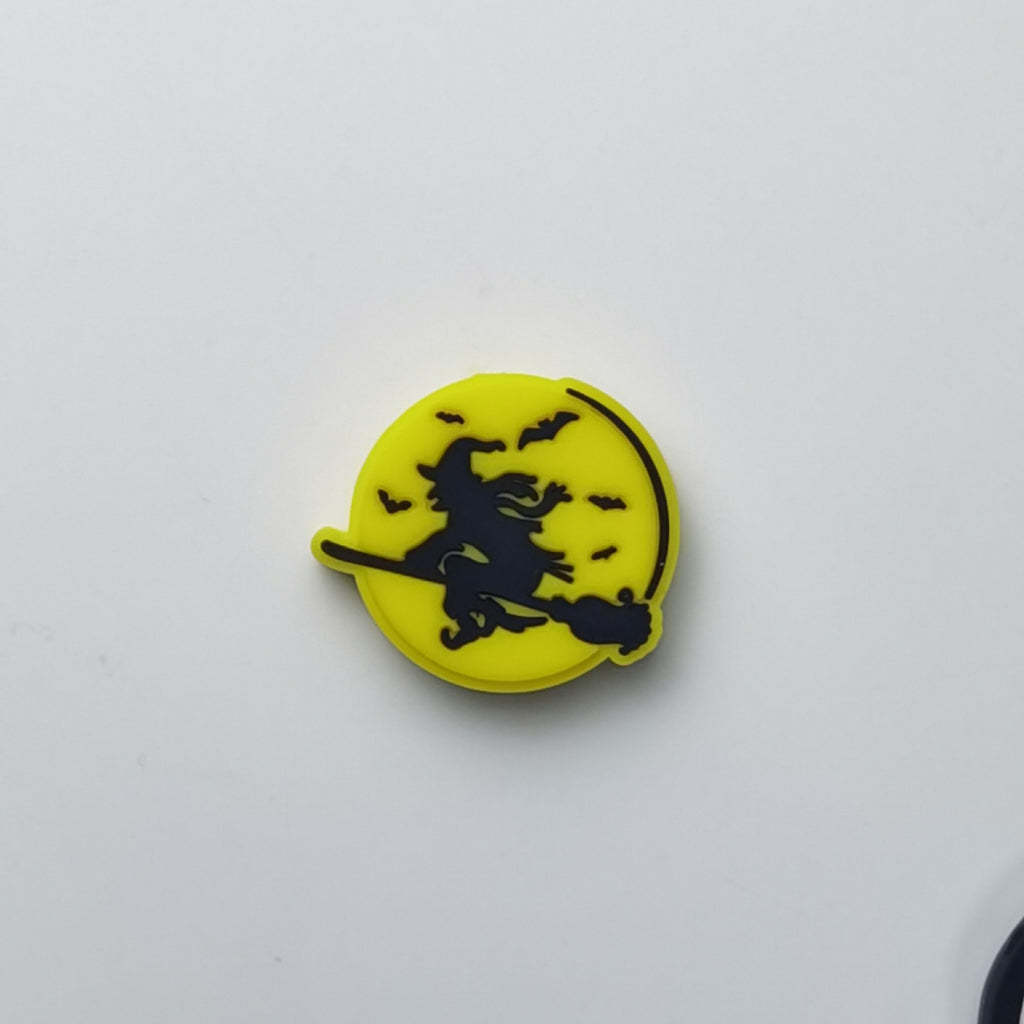 A small circular yellow accessory with a black silhouette of a witch riding a broomstick against a crescent moon and bats, shown on a plain light background.