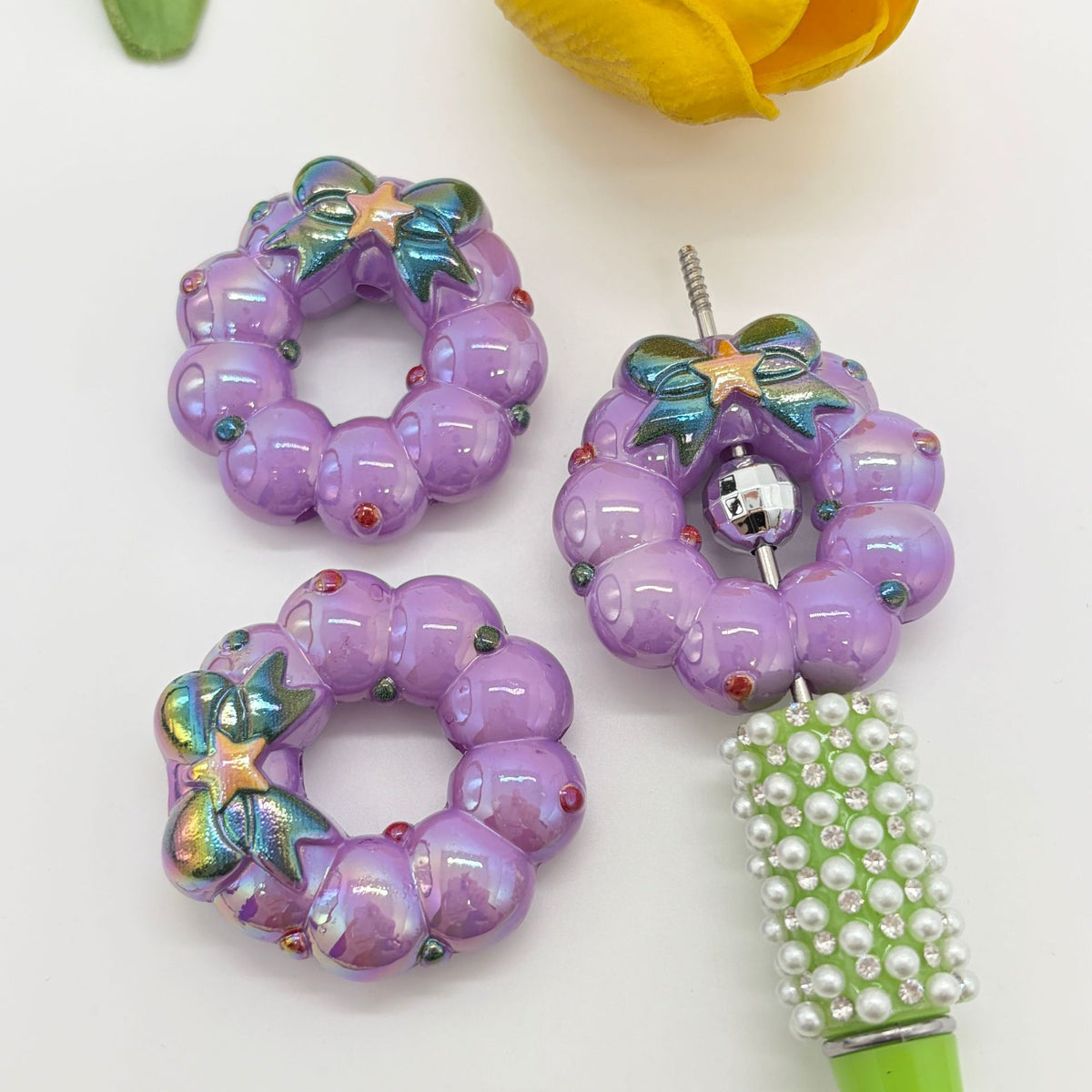 Three purple wreath-shaped acrylic focal beads with iridescent bow details on a white background