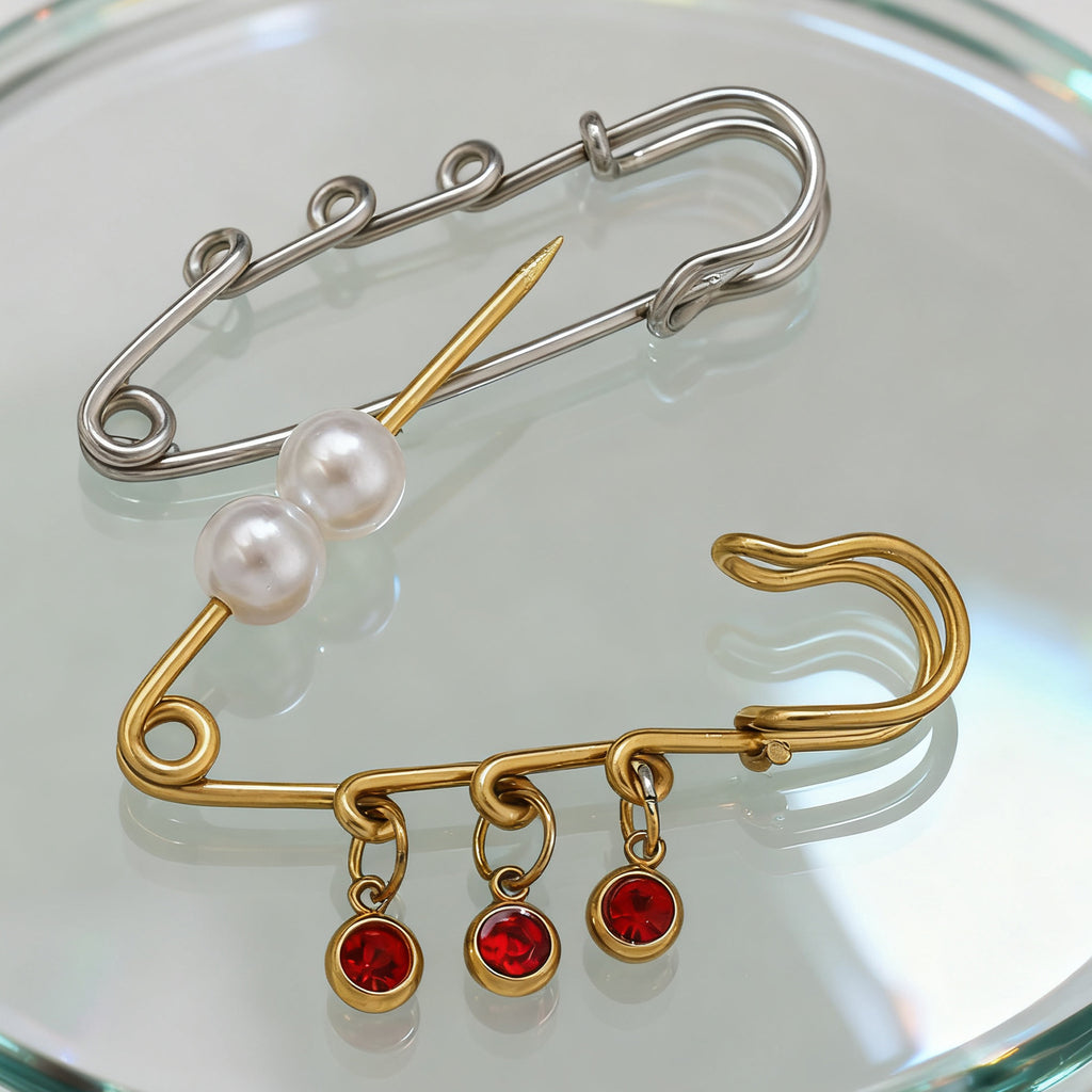 Silver safety-pin brooch with pearl beads and gold safety-pin brooch with red gemstone charms on a reflective surface