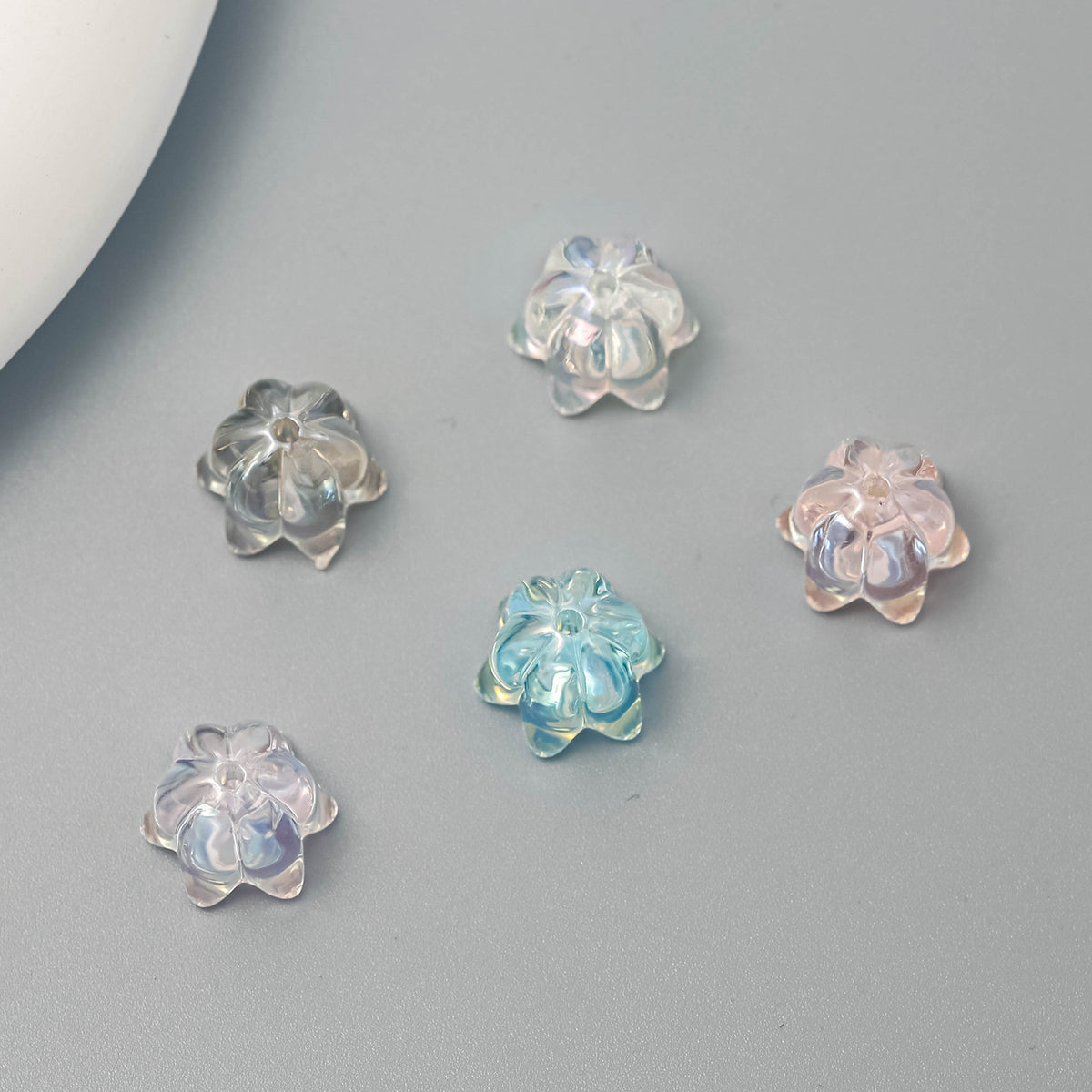 Five small translucent lily-shaped glitter beads in clear, light blue, pale pink and lavender on a smooth gray surface