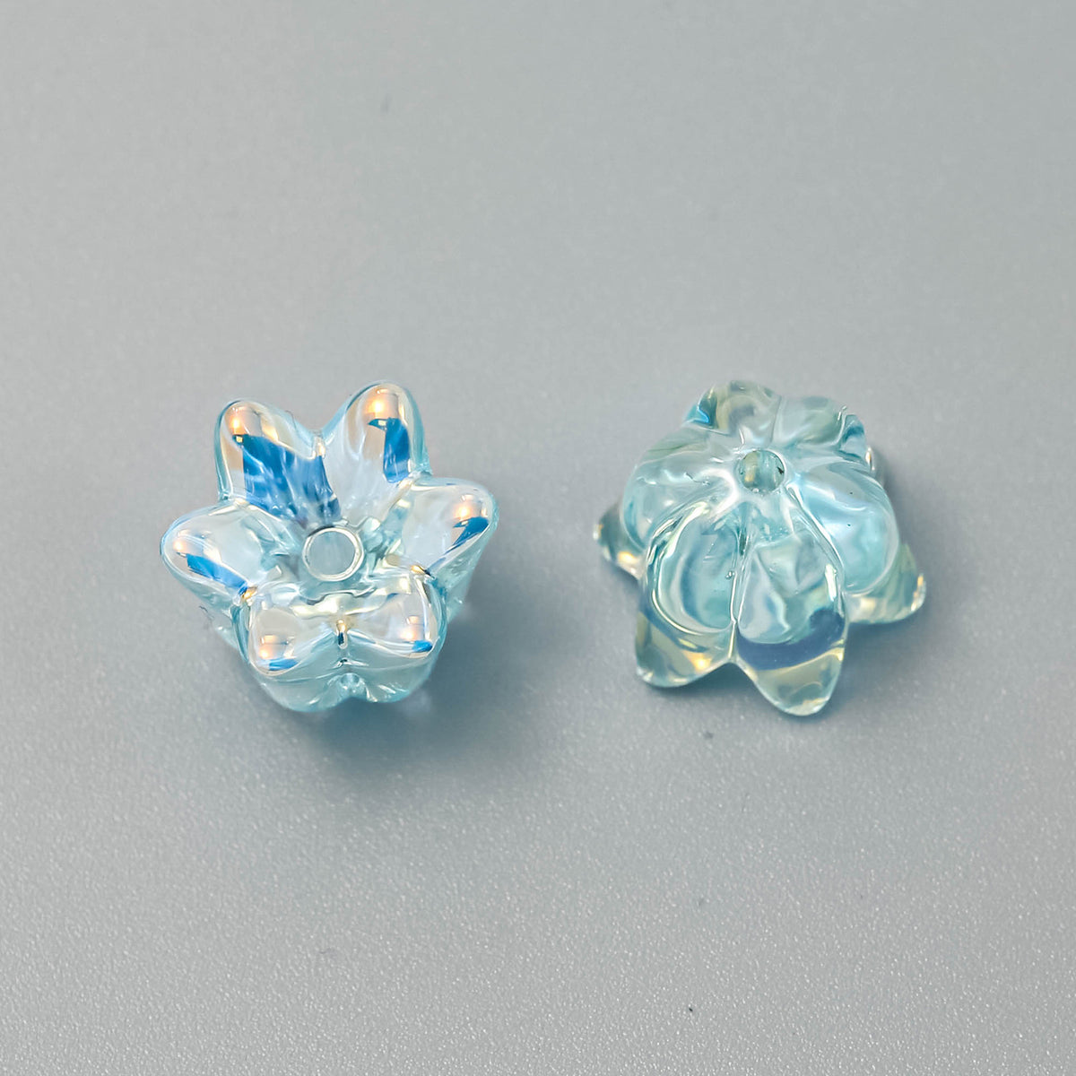 Two light blue iridescent lily-shaped beads, one showing front and one showing back, on a neutral gray background