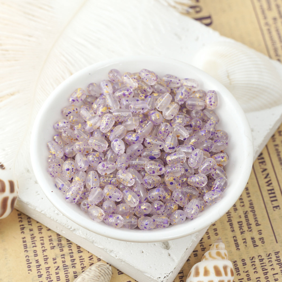 Translucent lavender glass teardrop beads speckled with purple and gold glitter in a white ceramic bowl on vintage newspaper.