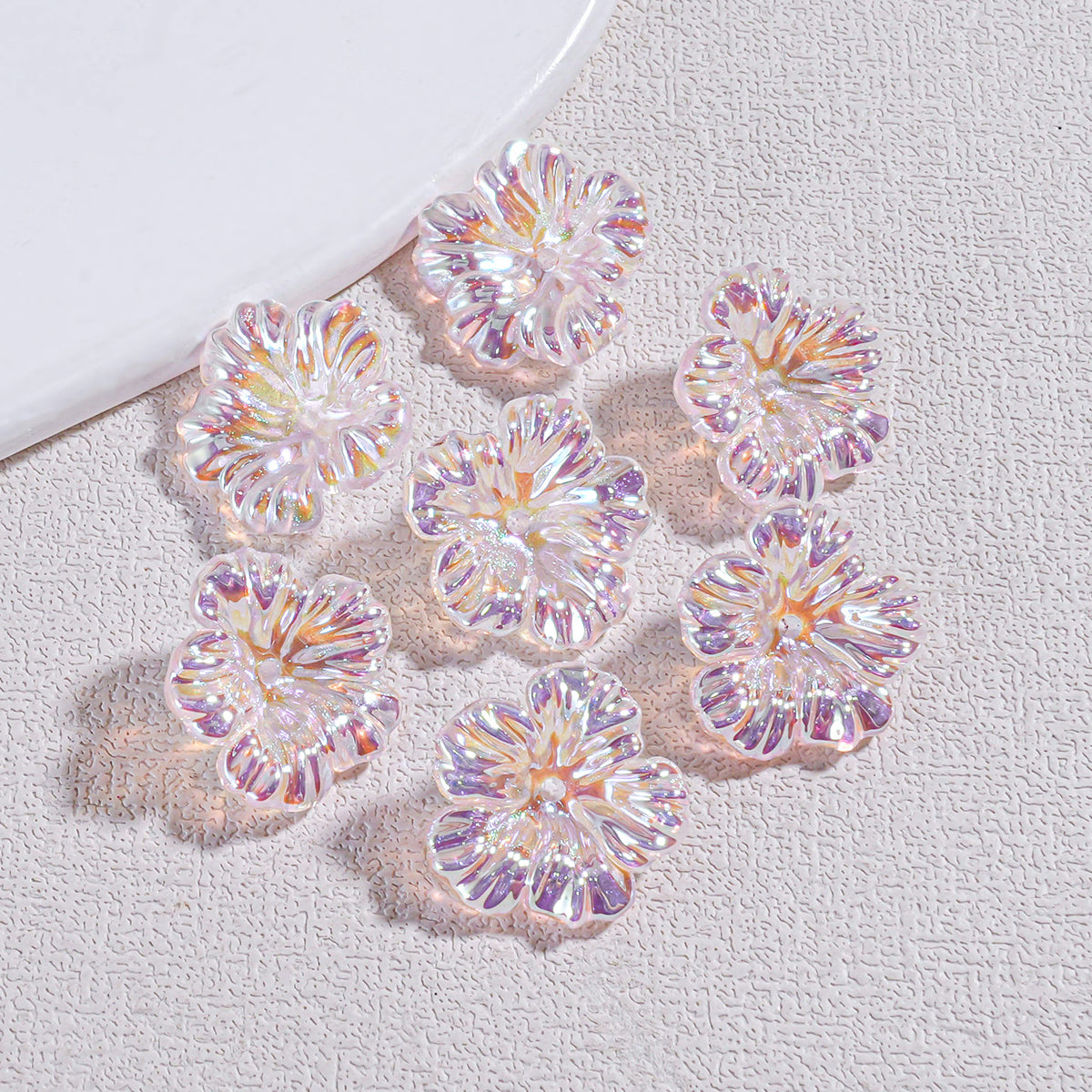 Seven iridescent holographic resin flower beads on a textured gray surface by a white plate edge
