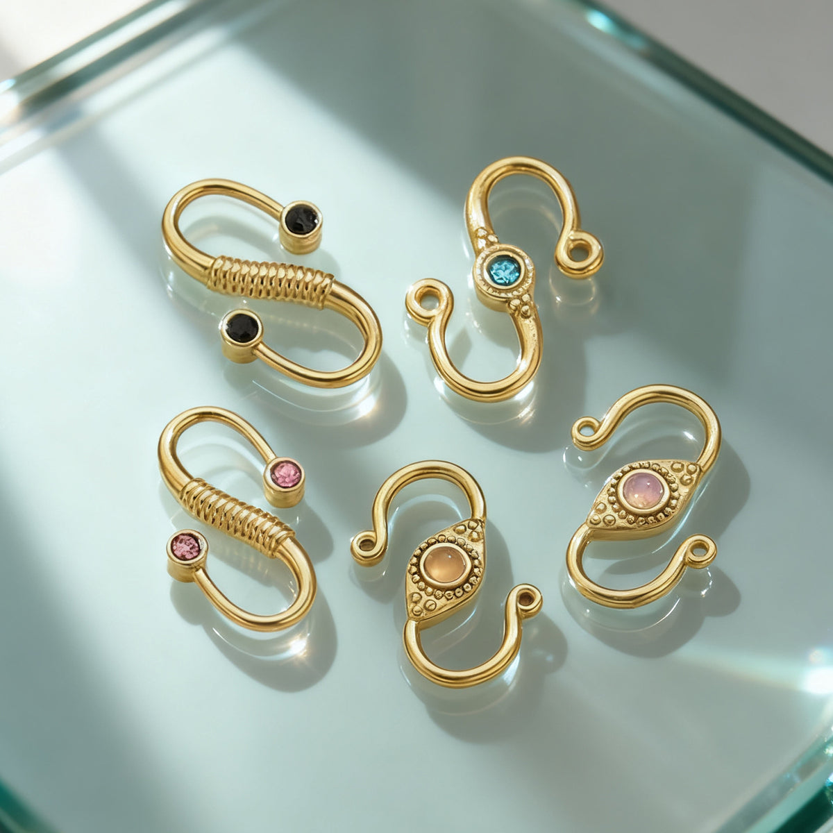 Five gold-tone S-hook jewelry clasps with colorful accent stones arranged on a light green glass surface