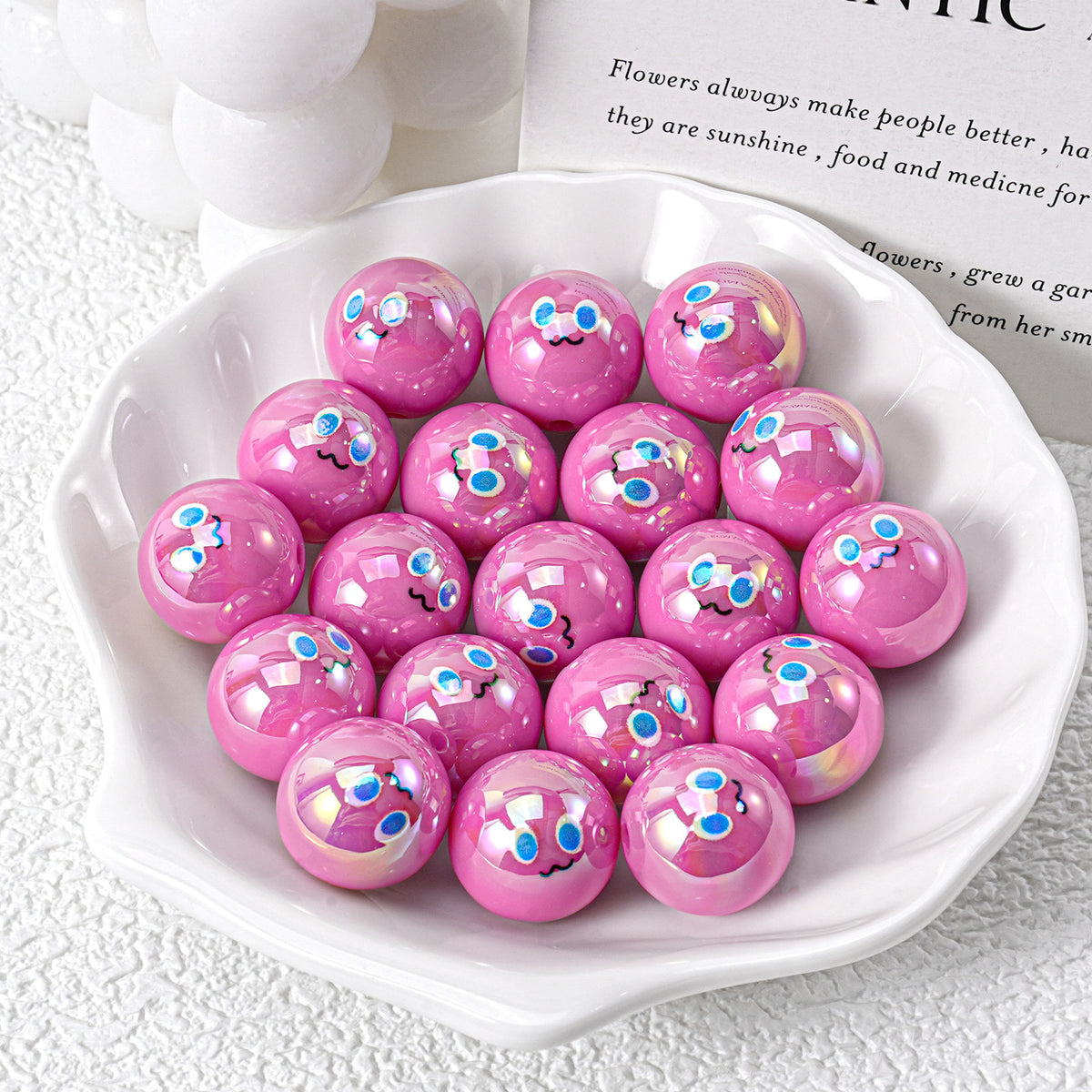 Glossy pink iridescent resin beads with cartoon faces in white ceramic dish on textured white surface