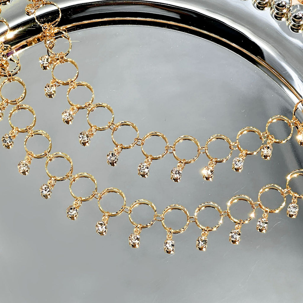A close-up of a gold-tone chain necklace with repeating circular links and small clear rhinestone drops, draped over a reflective silver surface.