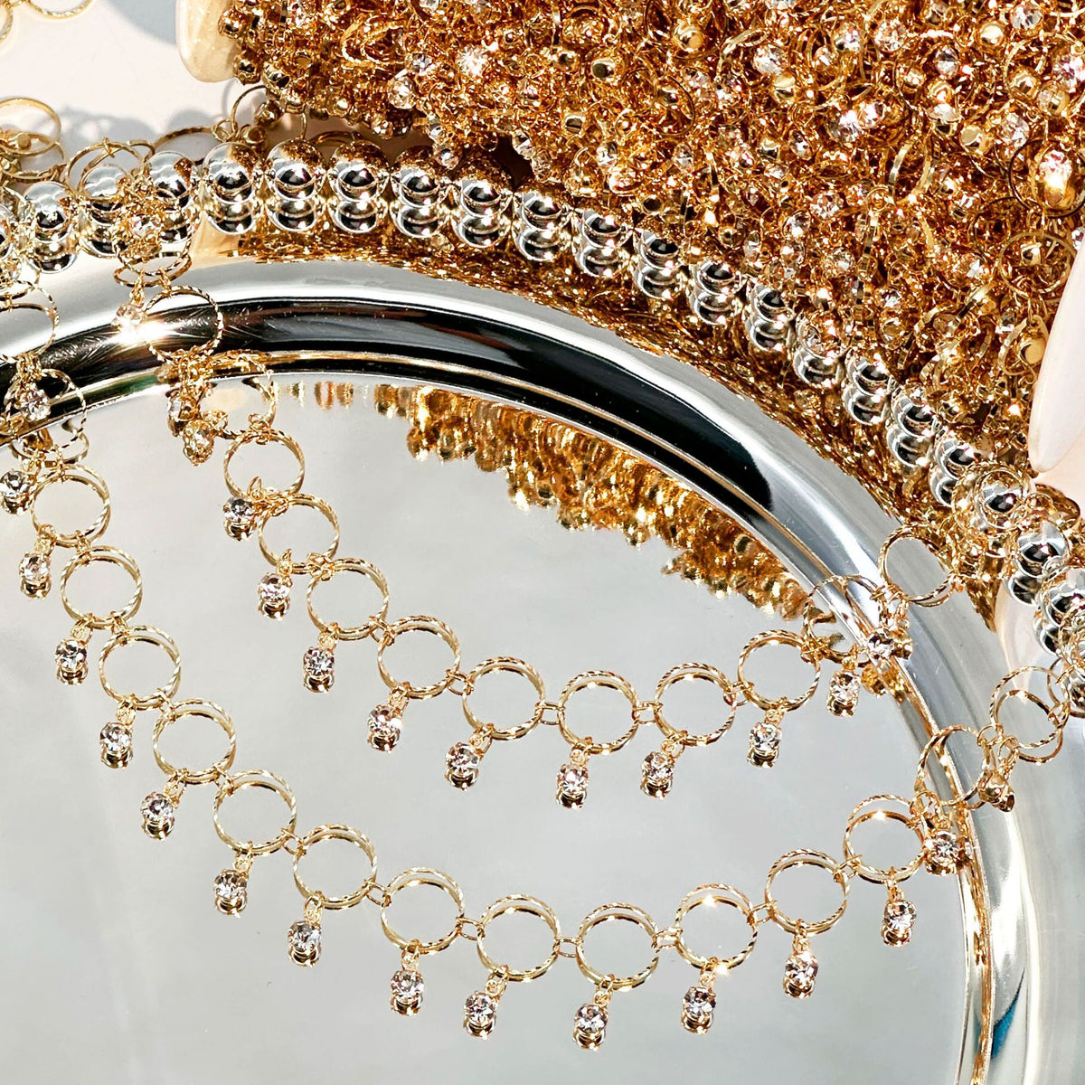 A close-up of a gold beaded fringe trim draped over a reflective silver tray, with a matching gold beaded fabric or accessory in the background.