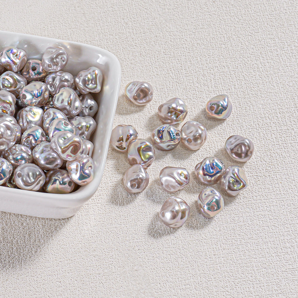 Iridescent silver-white baroque faux pearl beads in a white ceramic bowl with several scattered on a beige surface
