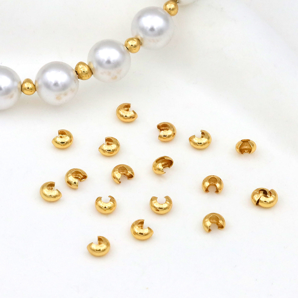 Gold C-shaped crimp bead covers scattered on white with a white pearl beaded bracelet in the corner