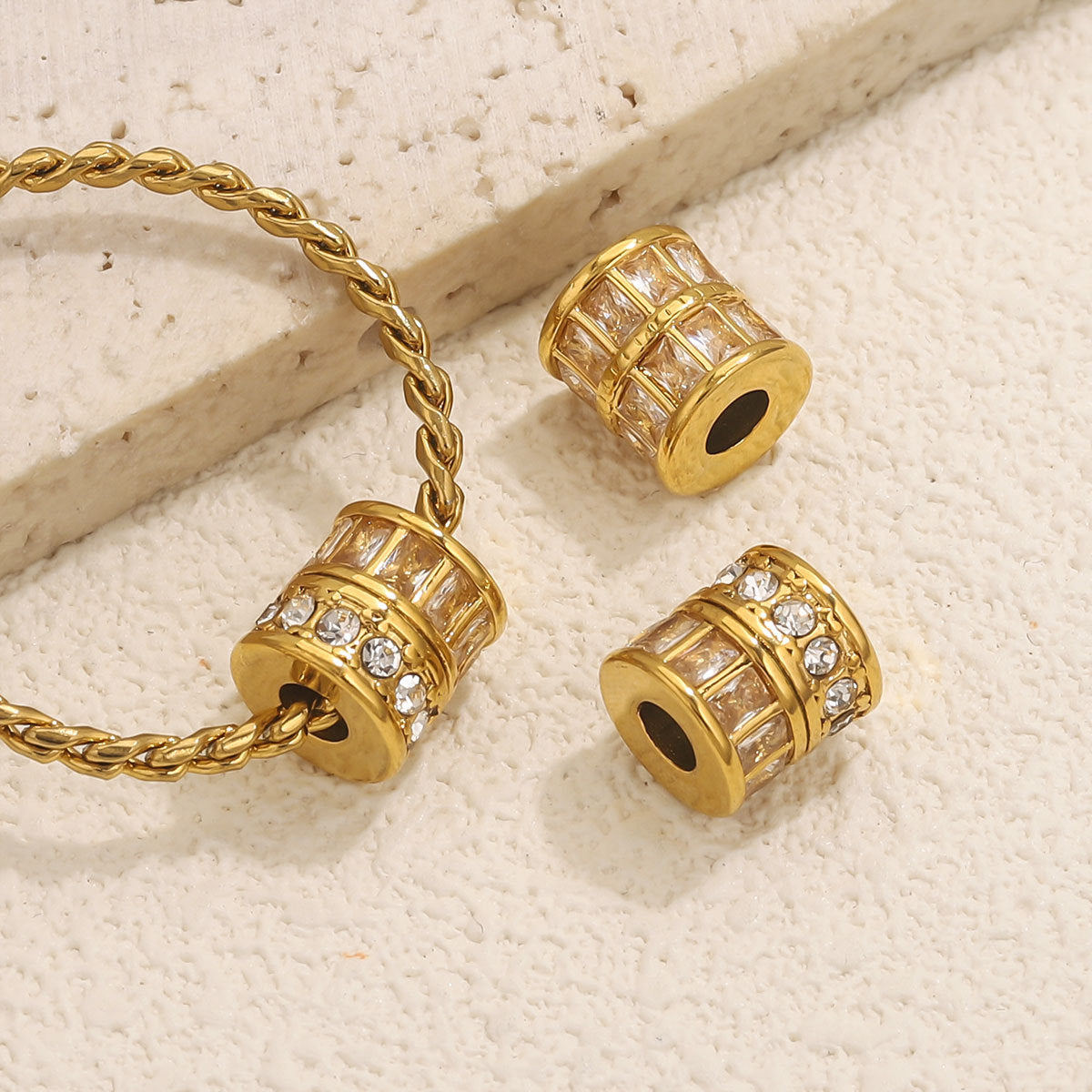 Three gold cylindrical charm beads with clear rhinestone accents, including one shown threaded on a gold chain and two displayed separately, on a textured cream surface.