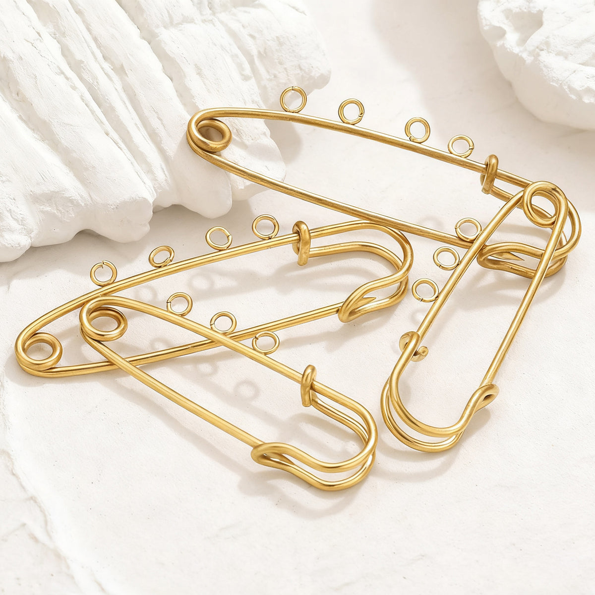 Two gold wire safety-pin style brooch pins with decorative loops on a textured white surface with soft shadows