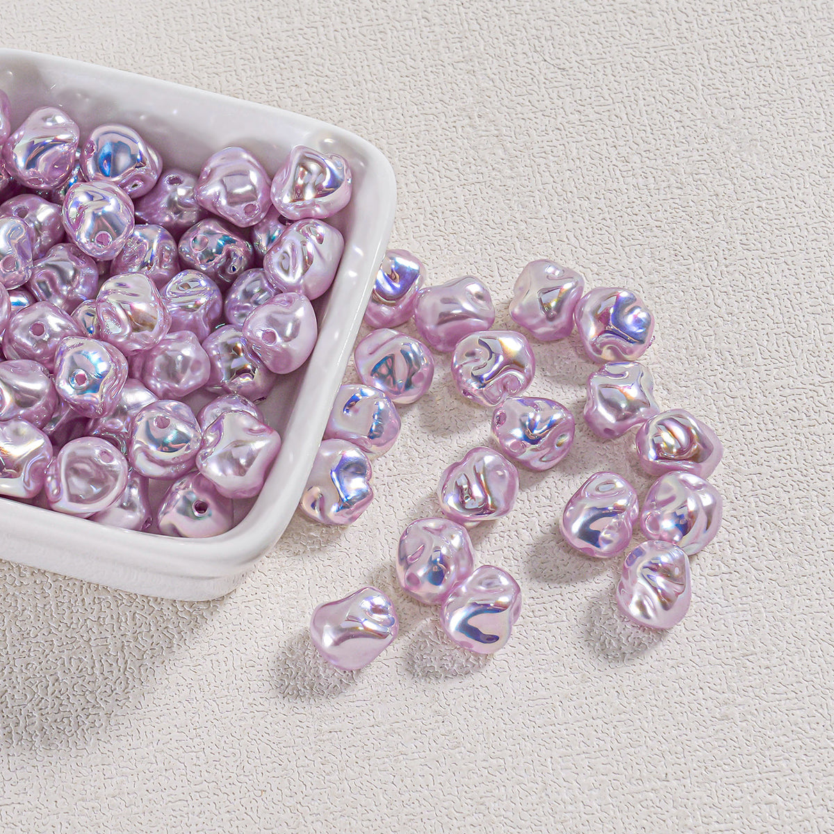 Shiny lavender iridescent baroque faux pearl beads in a white square dish with some spilled onto a beige surface