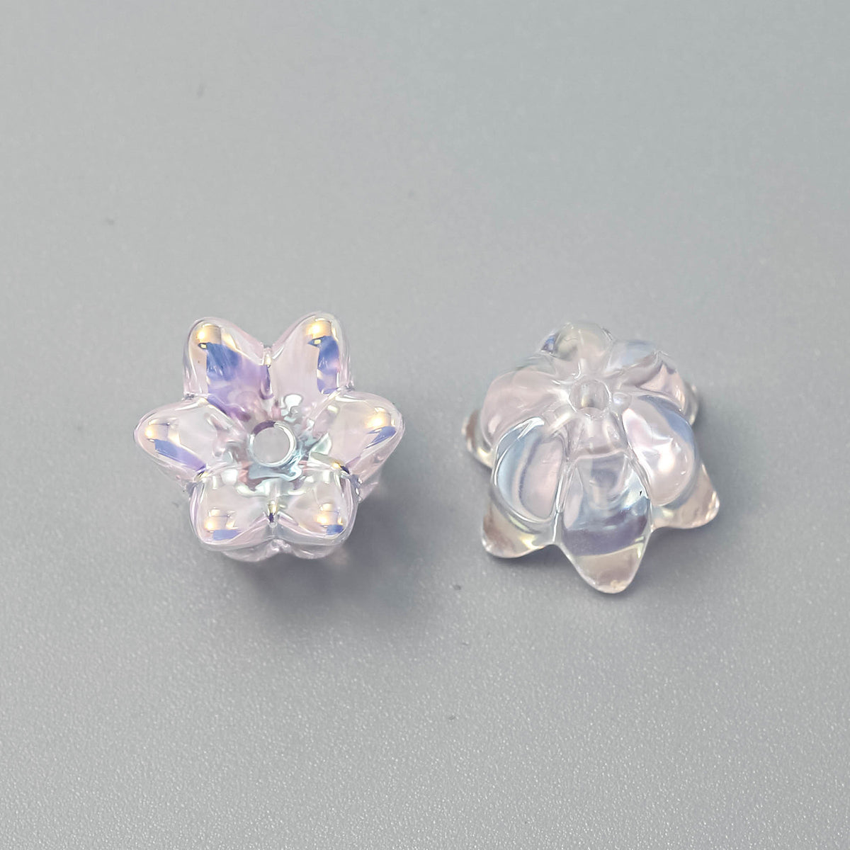 Two clear iridescent lily-shaped beads with holographic finish on a neutral gray background