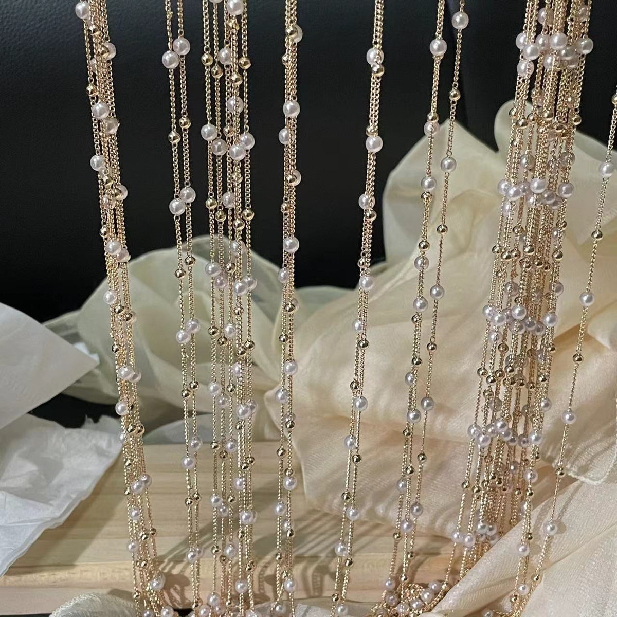 Gold-tone chains with white faux pearls hanging vertically over cream fabric backdrop