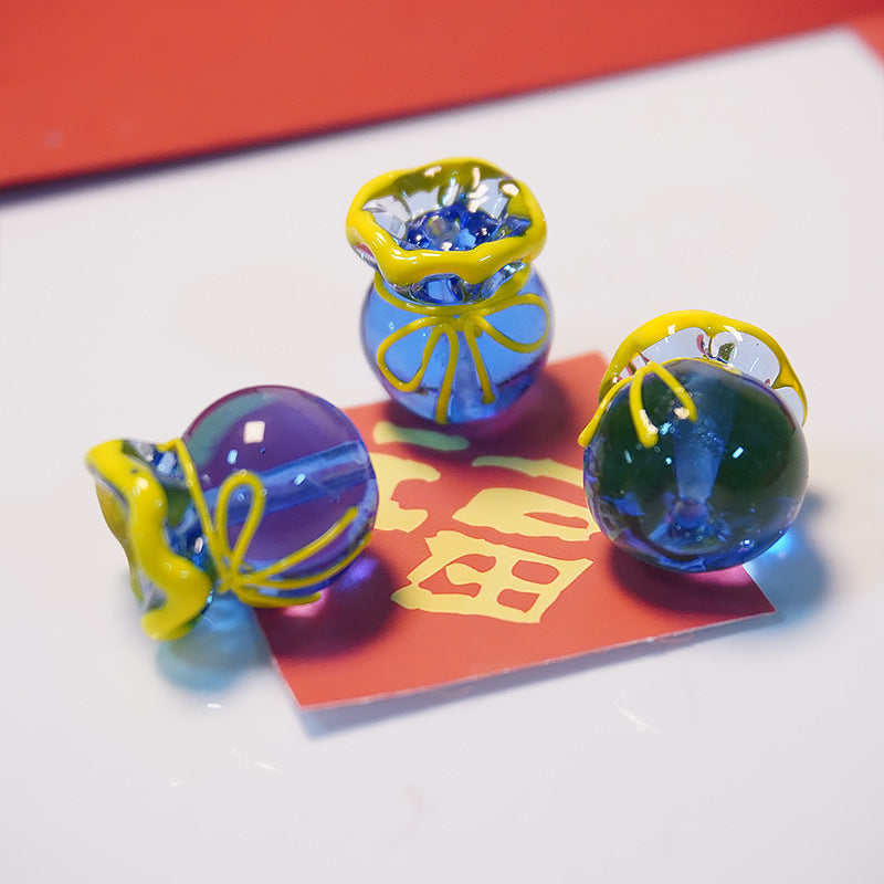 Three decorative glass ornaments shaped like money bags, featuring blue and green translucent bodies with yellow rope details, arranged on a white surface with a red card underneath.