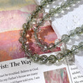 Strand of faceted clear beads with greenish-gray internal inclusions draped over printed paper and a white textured surface