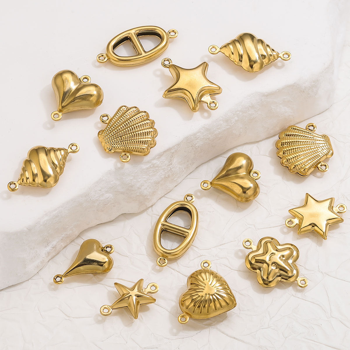 Gold-plated stainless steel double-hole hollow connector charms in heart, star, shell, and oval shapes on white fabric