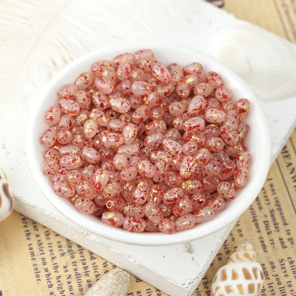 Translucent pink glass teardrop beads speckled with red and gold glitter in a white ceramic bowl on vintage newspaper.