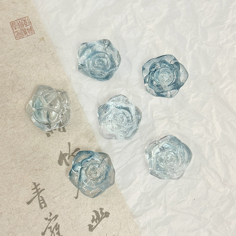 Five translucent light blue rose-shaped glass beads on a textured white surface with a beige calligraphy paper strip
