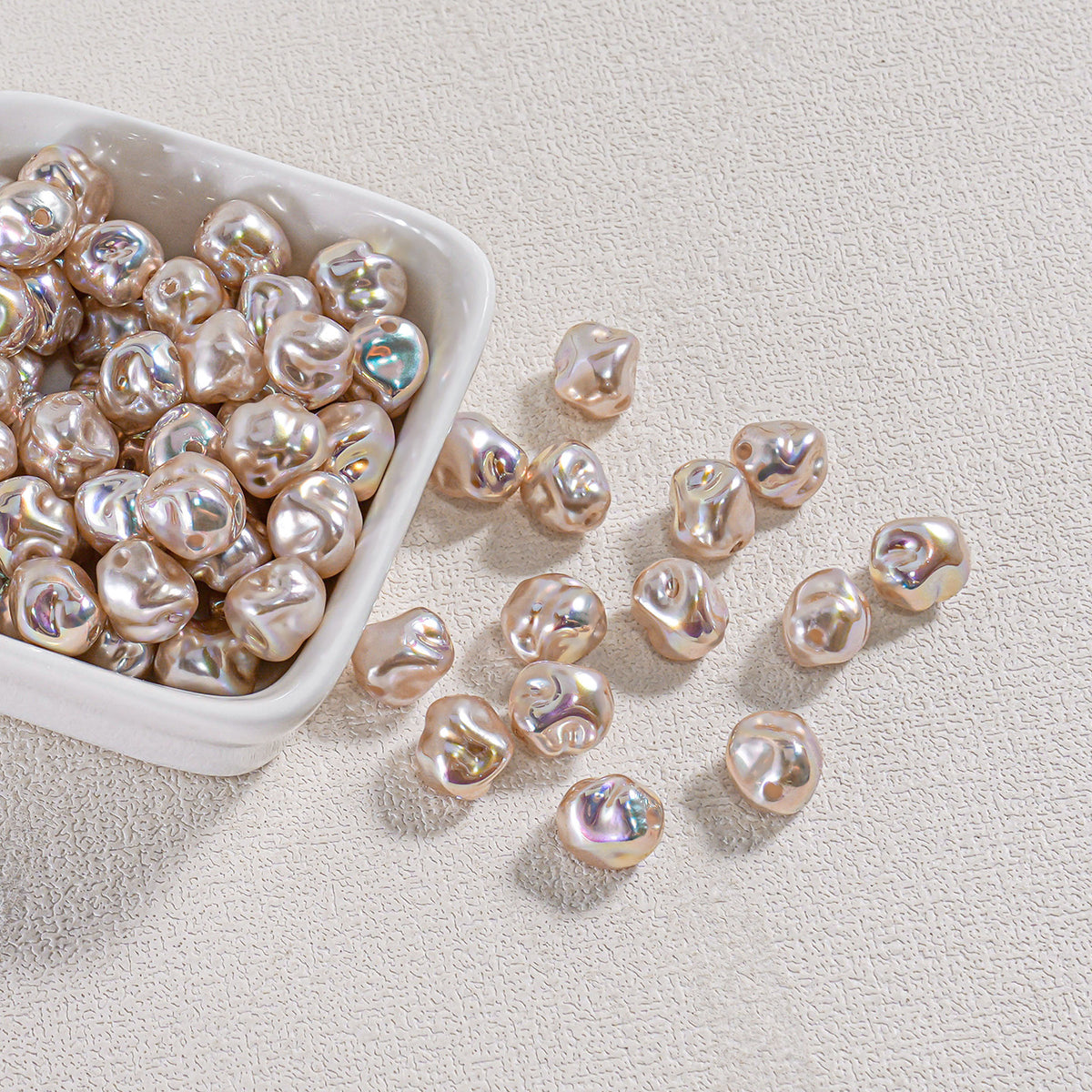 Iridescent champagne-gold baroque faux pearl beads in a white ceramic bowl with several scattered on a beige surface