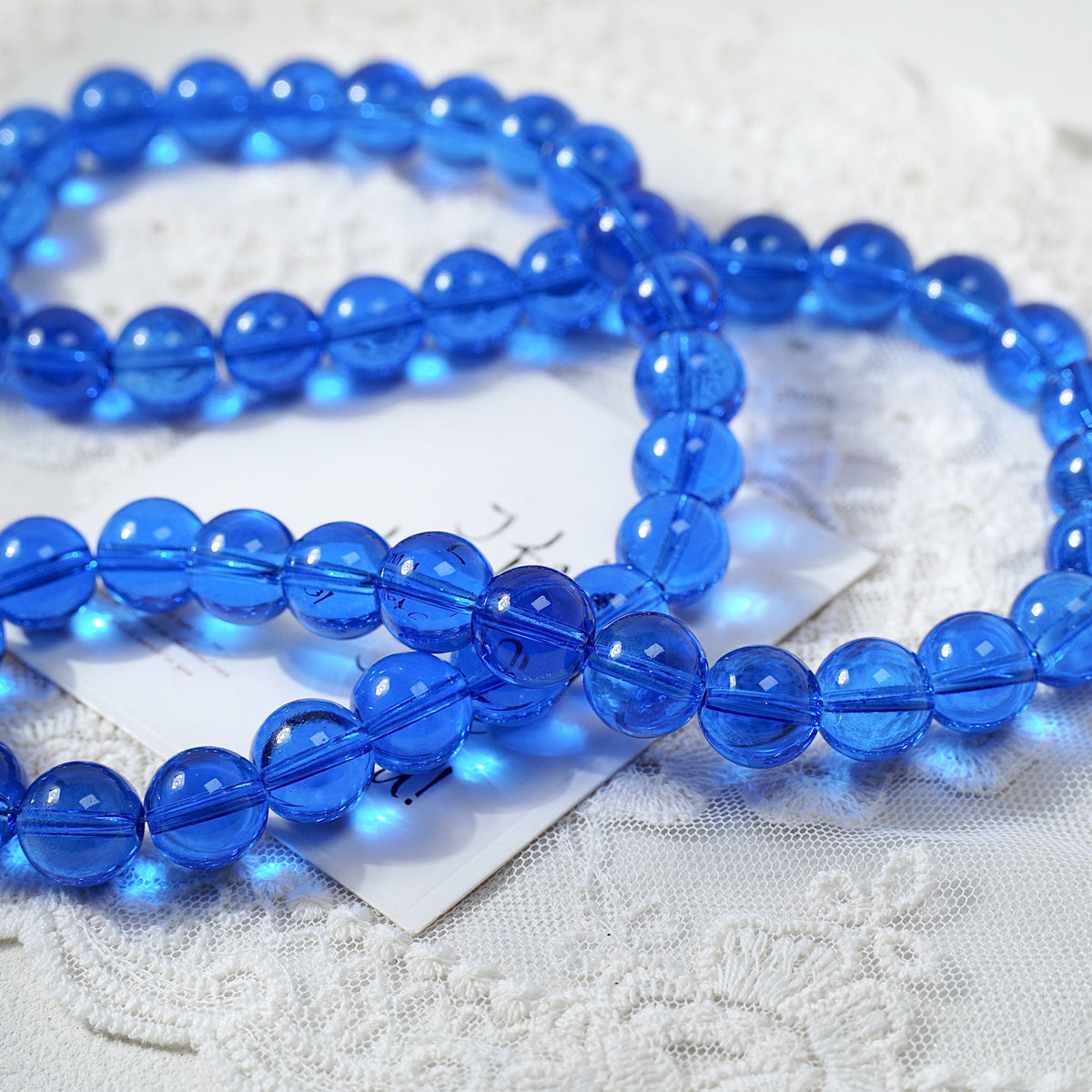 A strand of translucent blue glass beads arranged in loops on a white lace surface, with a small white tag