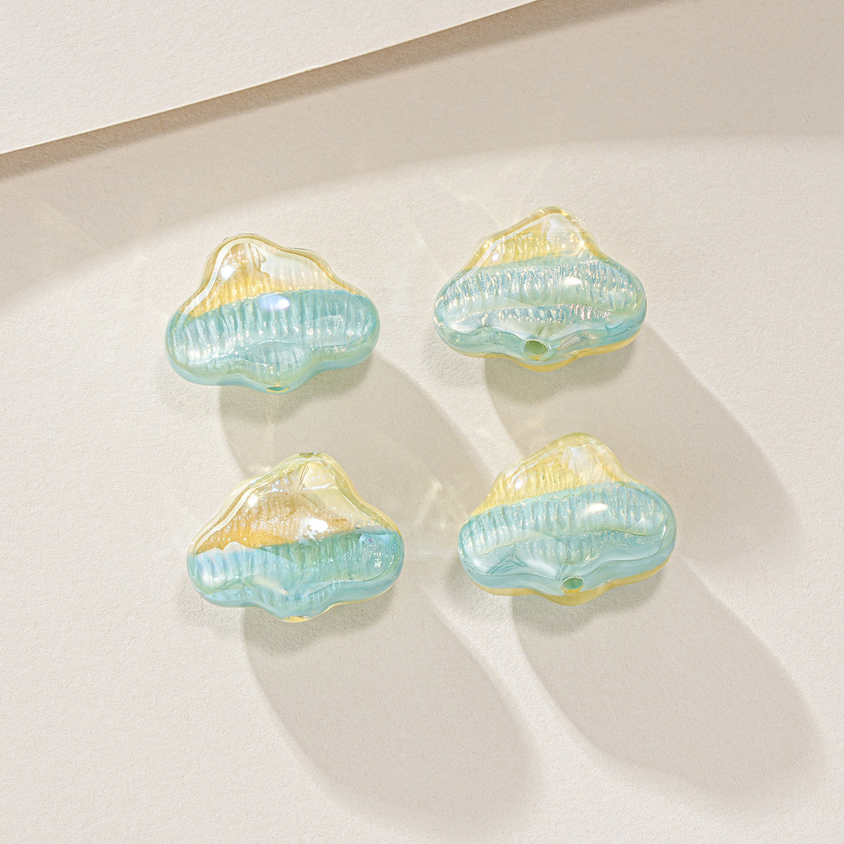 Four small cloud-shaped translucent beads in light blue and pale yellow, arranged on a warm off-white surface.