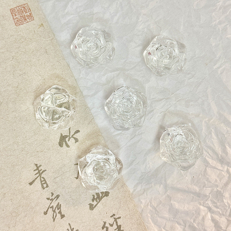 Five small clear glass rose-shaped beads on a textured white surface with a beige calligraphy paper strip