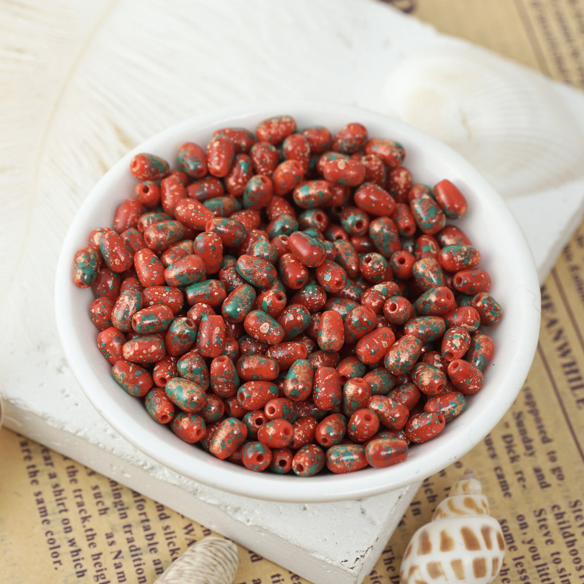 Red glass teardrop beads with teal speckles in a white ceramic bowl on vintage newspaper with seashells nearby.