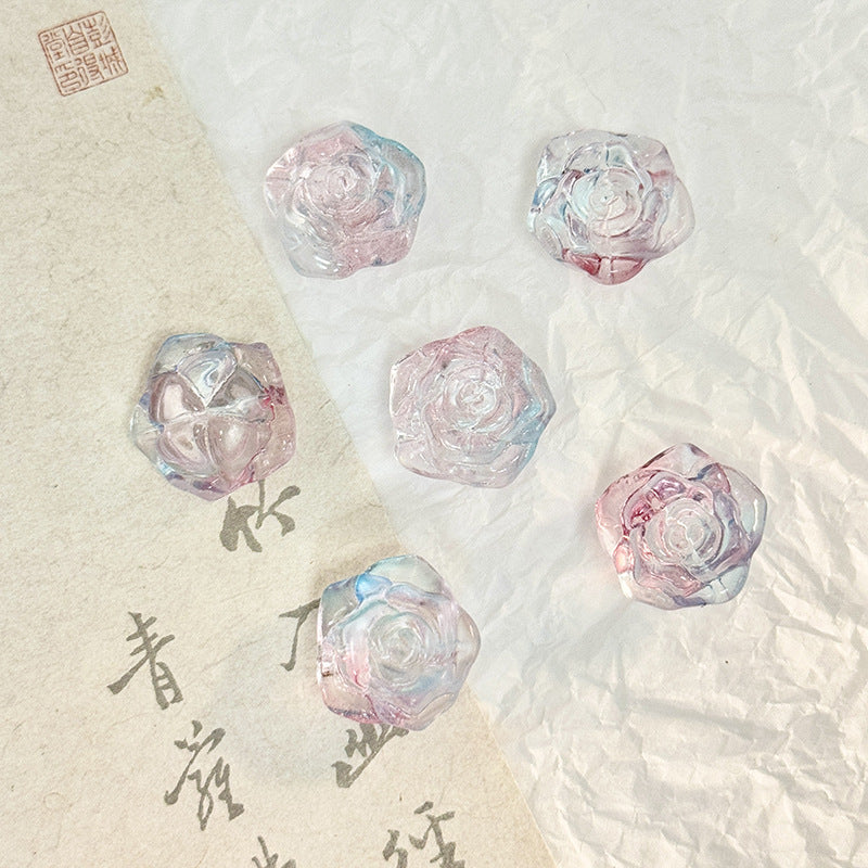 Five translucent iridescent rose-shaped glass beads in pink, blue and purple on a textured white surface